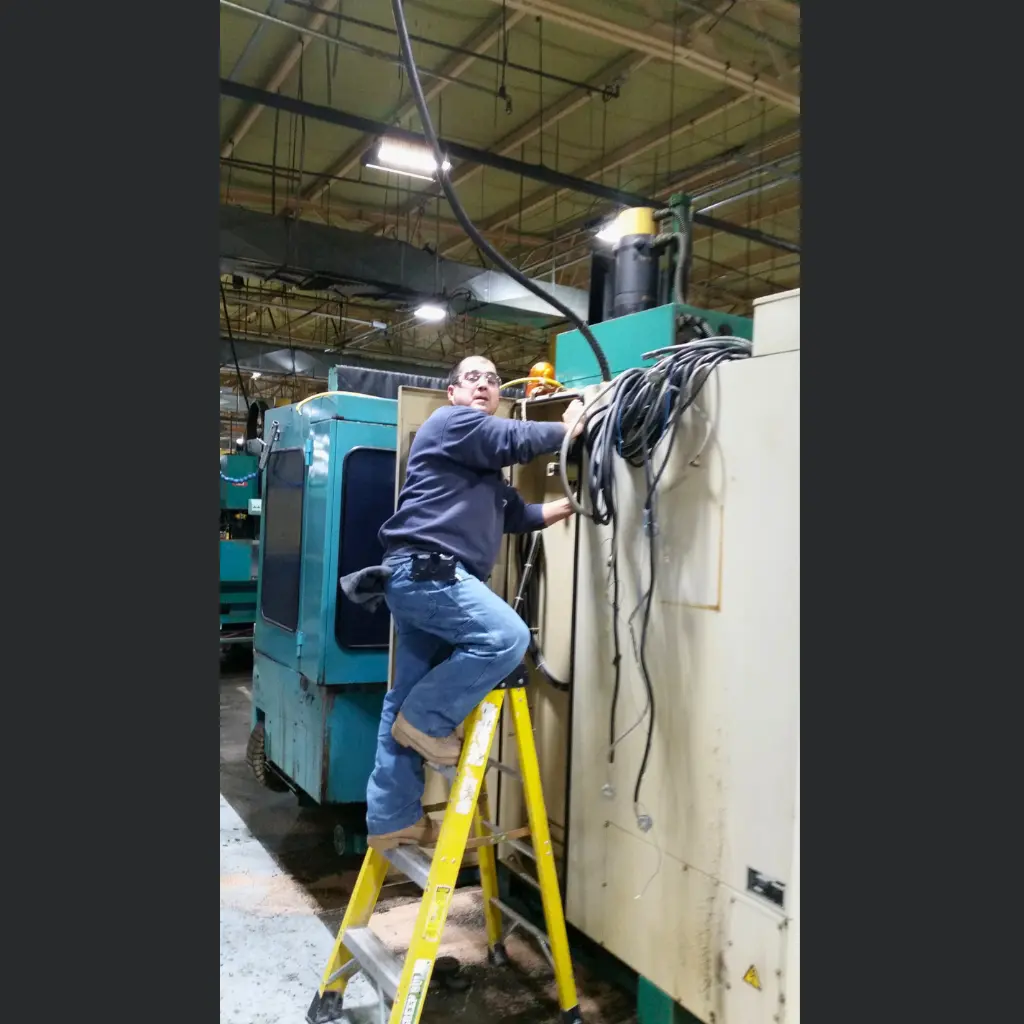 Electrician on a ladder performing electrical work beside industrial equipment